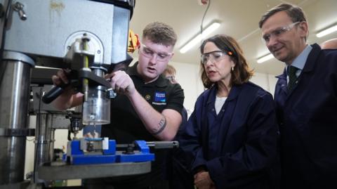 Work and Pensions Secretary Liz Kendall and Labour MP for Peterborough Andrew Pakes watch a young apprentice using a machine during a visit to Peterborough College. They are wearing safety goggles and overalls. 