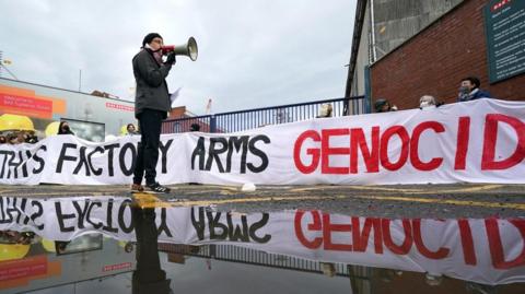 A protester holding a megaphone outside BAE Systems in Glasgow in May 2024. Behind him are red brick walls and the gates of the premises, and a long banner which reads "This factory arms genocide". The image is reflected in a puddle of water.