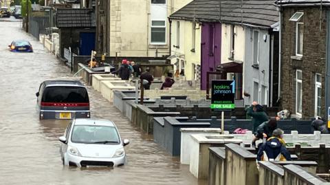 Flooded streets in Sion Street, Pontypridd. Three cars are deep in water in a street that looks more like a river. People wearing raincoats can be seen in the front gardens of terraced houses along the right hand side of the street. There is a pipe over the wall of one of the gardens being used to remove water from a property.