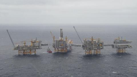 Four Equinor oil platforms in the North Sea. The grey platforms have yellow legs and cranes and a red and white boat is sailing between two of them. 