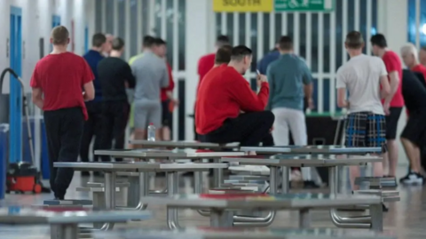 Prisoners wearing their own clothes and facing away from the camera congregate in a prison social space with tables and chairs