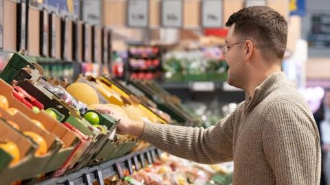 A man in a light brown jumper reaching for fruit in Lidl