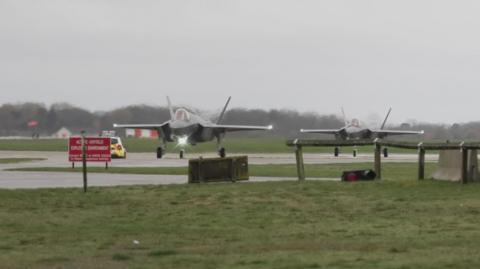 Two planes, which appear to be F-35As from the Joint Strike Fighter programme, located at RAF Lakenheath. They are located on a runway or taxiway, surrounded by grass lawns. A police car is visible. 