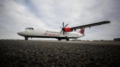 A Loganair aircraft on a runway with airport buildings in the distance behind it. It is a white plane with Loganair written on the side that has a red engine on the wing and a red tartan patter on the tail.