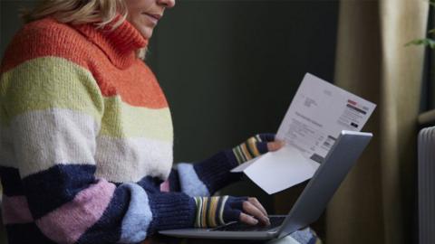 A woman sits in her home wearing a stripey jumper and gloves whilst reading a bill with her laptop on her lap