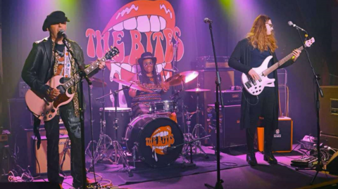 Three band members appear on a stage with two playing guitars and one man with a hat and dreadlocks on the drums. The two men standing are wearing mostly black. One has short hair and a hat on, the second long red/brown hair and glasses.