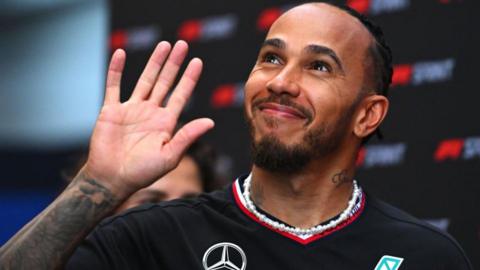 A smiling Lewis Hamilton looks up and waves