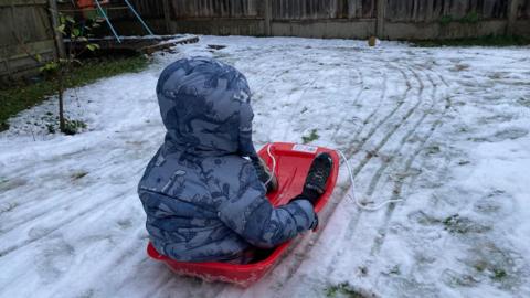 A child sits in a red plastic sledge in a snow-covered back garden. He is wearing a blue anorak. There is a swing frame and wooden fence in the distance.