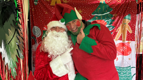 An elf, dressed in a red and green suit, gives the thumbs' up to Santa who is wearing his usual red and white costume, and sitting in a red Christmas grotto. 