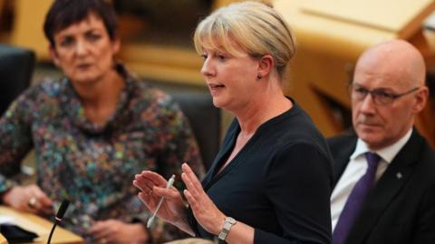 Shona Robison, with blonde hair and a black top, speaks in the Scottish Parliament with both hands raised in front of her. 