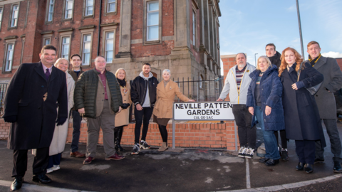 A group of 12 people in coats stood beside a street sign which reads Neville Patten Gardens with a large brick building standing behind them