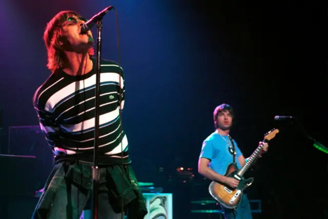 Noel and Liam Gallagher performing. Liam is in the foreground with a striped jumper on, Noel is behind playing a sunburst guitar.