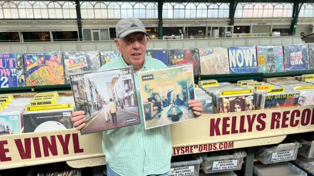 Oasis fan in blue striped shirt and jeans holding up two oasis vinyl albums (what's the story morning glory? and definitely maybe) in  a record store with kelly's record on sign and multiple rows of records stored behind him