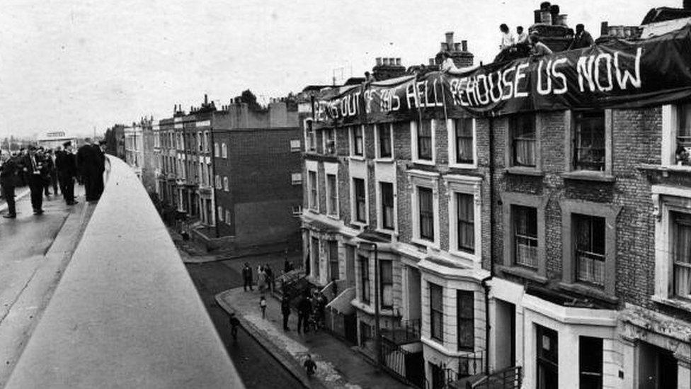 A banner saying 'Get Us Out Of This Hell Re-house Us Now' flies over a group of homes when the 'Westway' in London was opened.