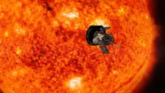 Художественное изображение космического аппарата Parker Solar Probe, пролетающего мимо Солнца, которое выглядит ярко-оранжевым, желтым, коричневым и черным и значительно превосходит по размерам космический аппарат на переднем плане