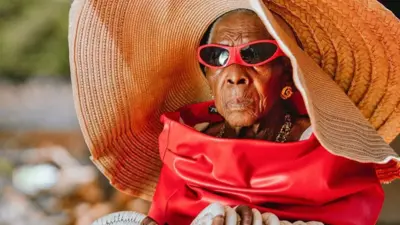 Margret Chola est glamour dans un chapeau en osier surdimensionné, une veste en cuir rouge, des bijoux en or et des lunettes de soleil rouges.