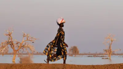 Une femme vêtue d'une robe noire imprimée d'ananas marche le long d'une digue en terre, un paquet sur la tête. De l'autre côté de la digue se trouvent plusieurs arbres entourés par les eaux de crue - Bentiu, Sud-Soudan