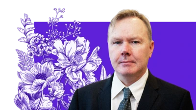 A man in a black suit with a white shirt and blue tie. He has short blond hair and is in front of a purple background with flowers