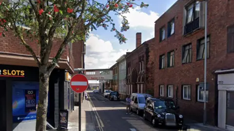 Google A city street with cars parked down one side and buildings on either side