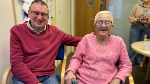 BBC Eddie and Maggie sit side by side in an office. They are both smiling broadly at the camera. He is wearing a red jumper and she is wearing a pink jumper.