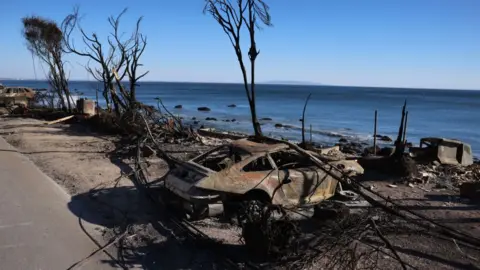 A burnt out car can be seen on the beach front in Malibu, with a clear view of the ocean visible behind it 