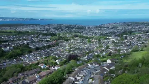 A drone photo of the area of Brixham which shows lots of houses, roads and some trees as well as the sea further away