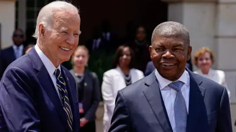 Joe Biden on the left smiles squinting into the sun as he shakes the hand of Joao Lourenco as they meet in Luanda.