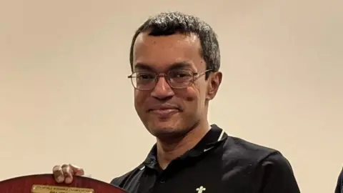 Harshan has short black hair, glasses, and a black sports shirt. 