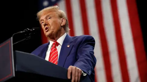 Donald Trump stands at a podium wearing a blue suit and red striped tie with an American flag in the background