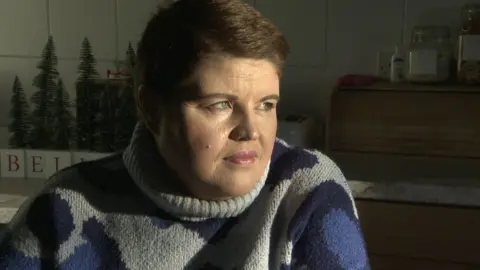 Emma - a woman with short brown hair has her head turned from the camera looking out the window as sun lights up her face. She is sitting down in a kitchen and is wearing a grey turtleneck jumper with large blue spots.