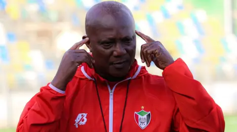 Kwesi Appiah, wearing a red tracksuit top with a Sudan FA logo on the left breast, points his index fingers to his temples