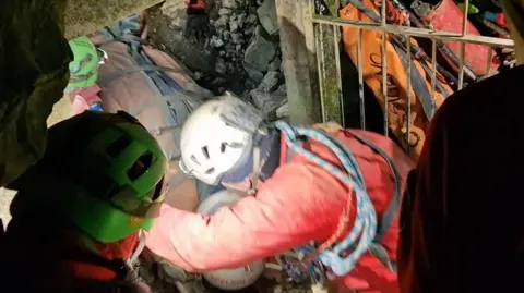 A woman strapped on to a stretcher, wearing a helmet, is pulled out from a cave in Italy, on the dirt ground beside rocks, through a grate, by rescuers wearing neon clothing, ropes and helmets 