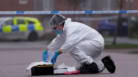 PA Media A forensic officer dressed in a white suit, hair net and blue gloves works with evidence equipment on a road within a police cordon. A police car can be seen in the distance.