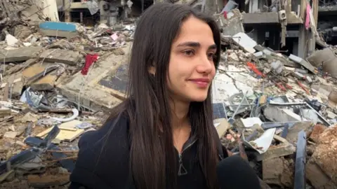 A young woman stands in front of rubble