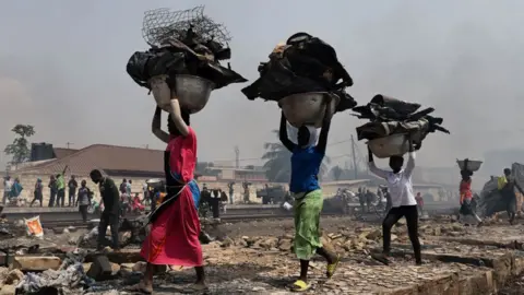 Three people carrying charred belongings in their heads walking through a fire-devastated landscape