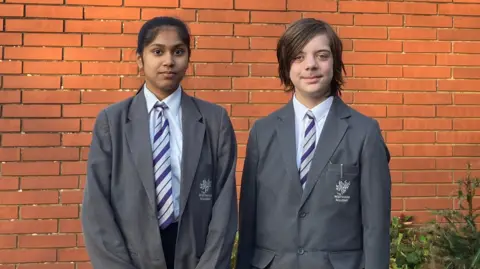 Students Nathan and Gargi looking at the camera. They are wearing school uniform made up a grey blazer and purple tie. They are standing in front of a brick wall and there are some flowers in the background. 