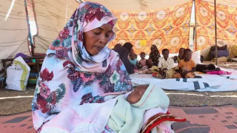 A woman in a headscarf, sitting on the floor in a large tent cradling a baby.