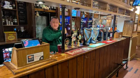 Terry Cole A man in a green jumper and short dark hair stood behind a bar with a number of pumps, pulling a pint. There is also a til on the bar and a number of bar towels