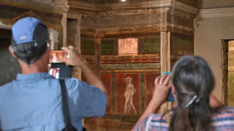 Two tourists wearing hats visit the Hall of Mysteries in the 'Villa of Mysteries' in Pompeii. They take photos on her phone of the wall paintings, which show images of Roman life