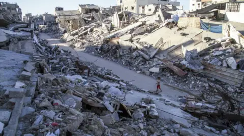 A wide shot of Jabalia, where a child stands on a road. On either side of the road is grey rubble.