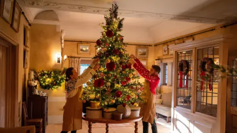 Two members of staff in aprons at Highgrove Gardens dress a Christmas tree, which is on a table, in a smaller room with red and gold decorations