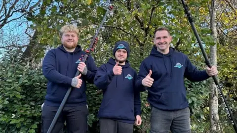 Three men in blue jumpers, holding various cleaning tools and putting their thumbs in the air, in front of a green hedge