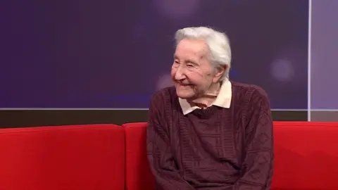 Edwin Rayner sits on a red sofa in the BBC South studio, there is a purple background. He is smiling, looking away from the camera, has white hair in a side parting and wears a mauve cable knit jumper with a nude coloured polo top underneath.