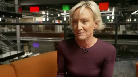 Ruth Dodsworth in the BBC studios in Cardiff. She is sitting on an orange sofa. She has cropped blonde hair and is wearing a fitted mauve top,