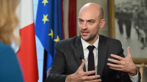 French Foreign Minister Jean-Noël Barrot speaking with the BBC's Laura Kuenssberg. He is wearing a dark grey suit, white shirt and dark red striped tie