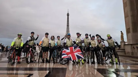 A team of cyclists stand in front of the Eiffel Tower with their bikes. One in the centre holds a Union Jack flag.