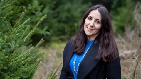 A woman standing among fir trees at a Christmas tree farm. She has long brown hair and is wearing a blue sweater with a blue lanyard and a black coat. She is looking at the camera and smiling. 