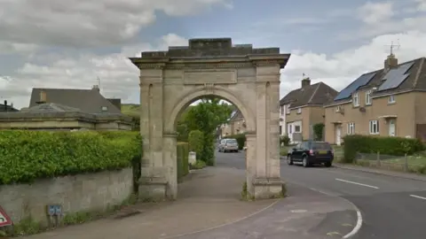 The Anti-Slavery Arch in Paganhill. It is a large rectangular stone archway on the site of a curved pavement, next to the road. There is a row of semi-detached houses on the opposite side of the road. Beside the archway is a low wall and hedge. 