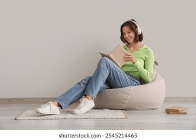 Mujer joven bonita en auriculares que leen el libro en la silla del beanbag en casa