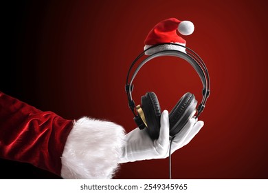 La mano de Santa sostiene los auriculares para escuchar la música de la Navidad decorada con el sombrero de Santa en un fondo aislado degradado rojo. Vista frontal